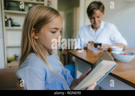 Mädchen mit Buch am Tisch zu Hause sitzen mit Mutter mit Handy im Hintergrund Stockfoto