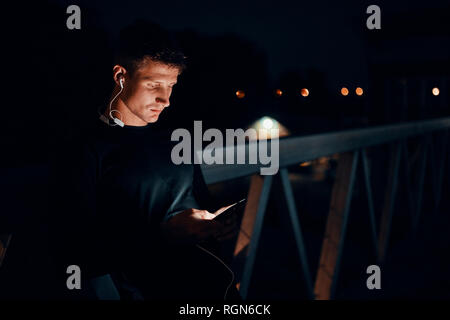 Sportlicher junger Mann mit Smartphone- und Ohrhörer im Freien bei Nacht Stockfoto