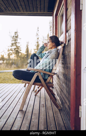 Schweden, Lappland, nachdenkliche junge Frau sitzt auf der Stuhl auf der Veranda mit Blick auf Abstand Stockfoto