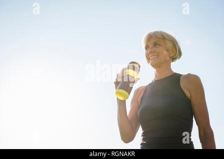 Lächelnd sportliche ältere Frau mit Flasche unter blauem Himmel Stockfoto