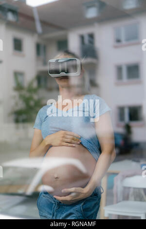 Schwangere Frau mit VR-Brille hinter Fensterglas Stockfoto