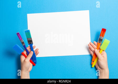 Kid Hände mit bunten Pinsel und leeres weißes Blatt Papier auf blauem Hintergrund Stockfoto