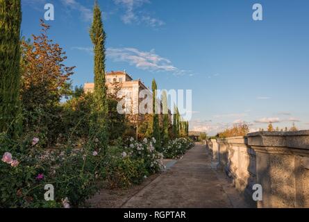 Ein Spanischer Garten mit Blumen am Rande eines Weges. Eine Wand ist auf der einen Seite und einem historischen Gebäude auf der anderen Seite. Ein blauer Himmel ist oben. Stockfoto