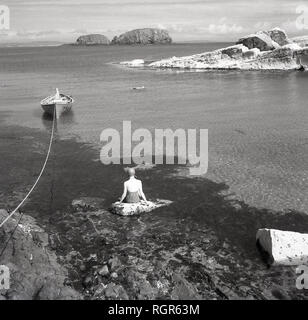 1950, historische, Nordirland, weiblichen Schwimmer schwimmen Hut tragen, Sitzen, Relaxen auf einem Felsen im seichten Meerwasser ein St einen geschützten Förde. Stockfoto