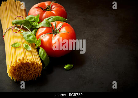 Bündel rohe Spaghetti unter Tomaten mit frischem Basilikum Blätter gegen den dunklen Hintergrund Stockfoto