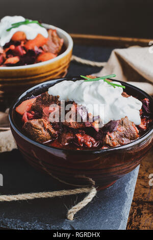 Zwei Schalen mit Borscht Suppe mit herzhaften, fleischigen Brocken von Rindfleisch, Wurzelgemüse, Kraut und Rüben. Mit saurer Sahne und Schnittlauch serviert, Stockfoto