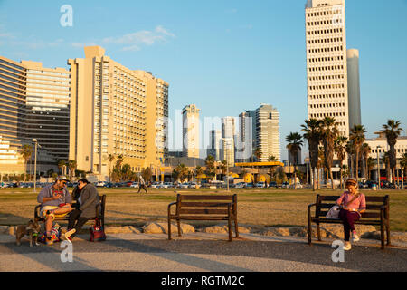 Tel Aviv, Israel - 23. Dezember 2018: Ein junges Paar Sonnenuntergang im Gegensatz zu der nächsten Bank sitzen einsam Mädchen mit Handy. Hinter ihnen ein Stockfoto