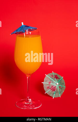 Orange Drink mit einem blauen Schirm in einem Glas Wein gegen eine feste rot hinterlegt. Ein grünes Dach liegt neben dem Glas. Stockfoto