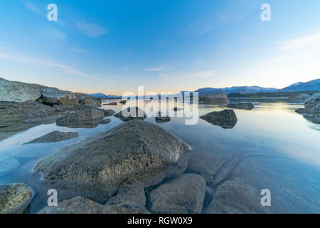 Großen Felsen in der Nähe der Kante Lake Tekapo als sunsises hinter die Berge in der Ferne und Vorland der Ruhe ruhige See Stockfoto