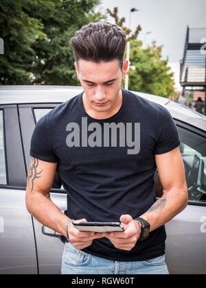 Hübscher junger Mann in Freizeitkleidung lehnte sich auf seinem Auto und mit Tablet-PC. Stockfoto