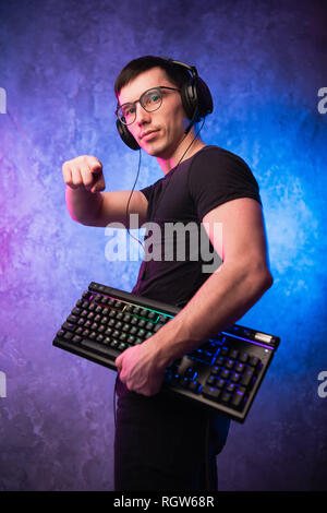 Junge schöne Gamer das Headset mit Mikrofon, halten Sie eine Tastatur und einen Zeigefinger an der Kamera auf Neon farbigen Hintergrund Stockfoto