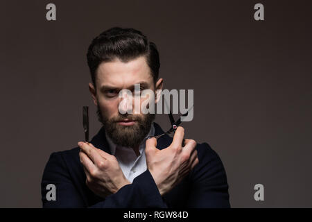 Bärtiger Mann kaukasischen Hipster mit Schnurrbart, Schere und Rasiermesser auf grauem Hintergrund. Mens Haarschnitt, rasieren. Männliche im barbershop. Stockfoto