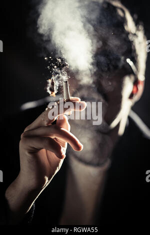 Junge Kerl in Brillen mit brennender Zigarette in der Hand zwischen den Dunst in der Dunkelheit Stockfoto