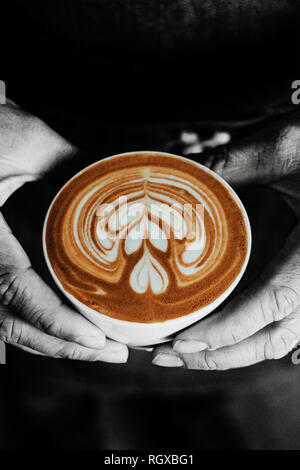 Eine Tasse Kaffee mit einem Herzen latte Schaum Kunst auf der Oberseite, helf von männlichen Händen, in Schwarz und Weiß, Monochrom. Stockfoto