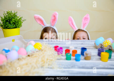 Neugierige kleine Mädchen in Hasenohren spähen hinter Tisch mit leckeren Ostereiern und Kamera mit Überraschung. Schwerpunkt Kinder Stockfoto