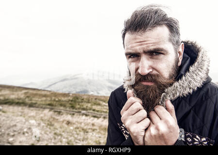 Bärtiger Mann, langen Bart, brutale kaukasischen Hipster mit Schnurrbart auf ernstes Gesicht, unrasierten Kerl mit stilvollen Haare, bärte Haarschnitt kauern von kalt auf dem Berg oben an bewölkten Himmel, Platz kopieren Stockfoto