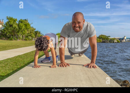 Ein Vater lächelt als er zeigt seinem Sohn, wie Liegestütze auf einer ufermauer am Intercoastal zu tun. Der Junge schaut Vati ihn lehren für eine gute Gesundheit. Stockfoto