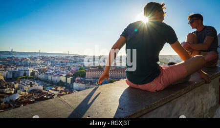 Zwei Männer genießen den Sonnenuntergang mit Stadtblick vom Miradouro da Graça, Lissabon, Portugal, mit rückseitig beleuchtetem Sonnenlicht Stockfoto