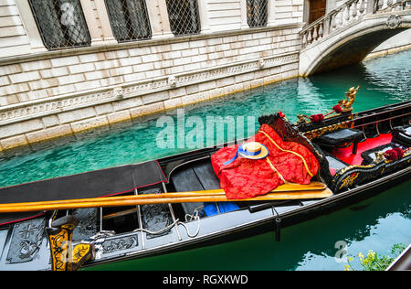 Ein Fisch schwimmt unter der Venezianischen Gondel mit Rudern, gondoliere Hut, gestreiftes Hemd und roter Decke, wie es unbesetzt in einem bunten Kanal in Venedig Italien sitzt Stockfoto