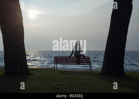 Frau sitzen auf einer Bank an der Uferpromenade zwischen zwei Bäumen in Ascona, Schweiz. Stockfoto
