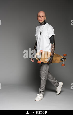 Stattliche tätowierten Mann im weißen T-Shirt mit Longboard und Kamera auf Grau Stockfoto