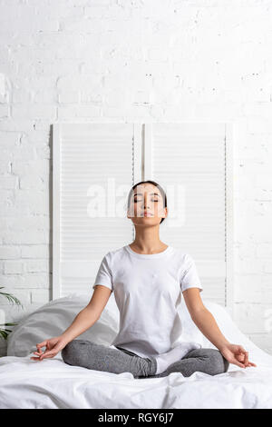 Asiatische Frau im weißen T-Shirt und leggings Grau sitzen in Lotus im Bett stellen Stockfoto
