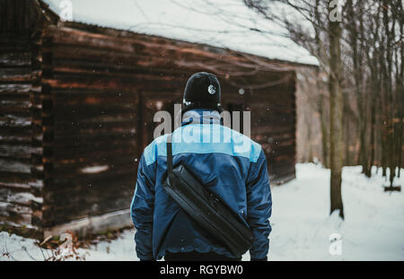 Foto der Abenteurer (Wanderer) im verschneiten Wald vor der verlassenen Hütte (Blick von seinem Rücken). Wanderer Mann in der blauen Jacke steht in den Bergen Stockfoto