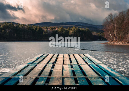 Mole (Pier) auf dem See. Hölzerne Brücke in Wald im Winter mit Blau gefrorenen See. See zum Angeln mit Pier. Dunklen See (ICE) mit Hügeln Stockfoto