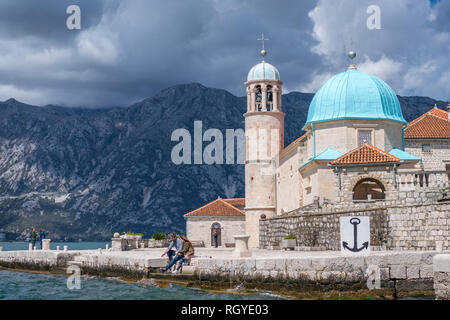 Perast, Montenegro - April 2018: Paar ruht auf dem Ufer vor dem Glockenturm der Basilika Unserer Lieben Frau von den Felsen in die Bucht von Kotor Stockfoto