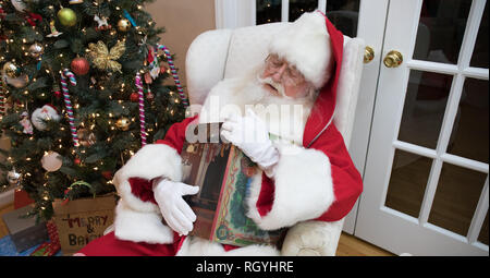 Santa Claus schlafend in einem weißen Stuhl hält ein Buch - Weihnachten neben einem Weihnachtsbaum. Stockfoto