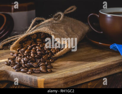 Verschüttete Kaffeebohnen aus Sackleinen Tasche auf einem Holzbrett mit Tasse Kaffee und Zucker Schale im Hintergrund in einer dunklen Umgebung Stockfoto
