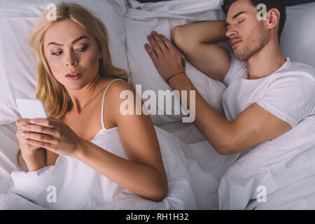 Blick von oben auf die Verdächtige junge Frau mit Smartphone und Freund schlafen im Bett Stockfoto