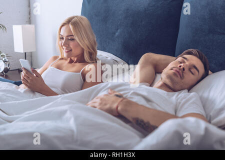 Hübscher junger Mann schlafen und lächelnde Frau mit im Bett smartphone