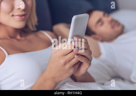 7/8 Schuß von lächelnden jungen Frau mit Smartphone, während Mann schlafen im Bett Stockfoto