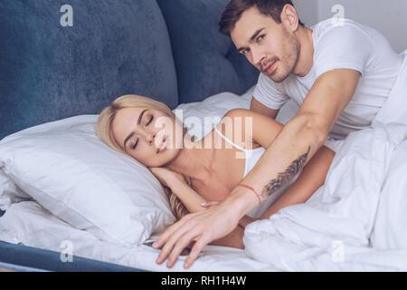 Junger Mann erreichen für Smartphone und die Kamera während der Freundin schlafen im Bett, Misstrauen Konzept Stockfoto