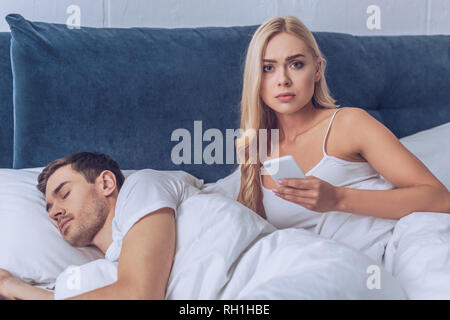 Unglückliche junge Frau mit Smartphone und Suchen an der Kamera, während Freund schlafen im Bett, Misstrauen Konzept Stockfoto