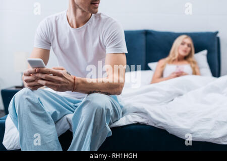 Der junge Mann im Schlafanzug mit Smartphone 7/8 während eifersüchtige Frau im Bett hinter Lügen Stockfoto