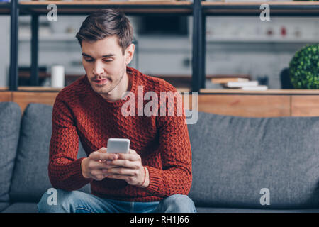 Nachdenklicher junger Mann sitzt auf einem Sofa und sie ihr Smartphone zu Hause Stockfoto