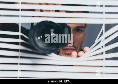 Junger Mann mit Kamera und Kamera durch Jalousien, Misstrauen Konzept Stockfoto