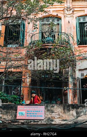 Hanoi, Vietnam, 12.20.18: Das Leben auf der Straße in Hanoi. Alte Dame auf einem Balkon in einem alten Gebäude. Stockfoto