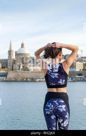 Rückansicht einer Frau, die ihre Haare einstellen, bevor Sie draussen trainieren. Valletta Malta Hintergrund. Stockfoto