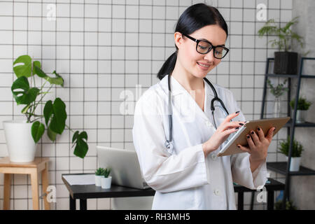 Weibliche brunette Arzt trägt eine Brille mit Tablet, intelligente und schöne Frau mit der Gadget und Stethoskop im weißen Büro Hintergrund. Stockfoto