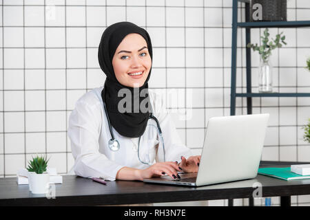 Wissenschaftlerin sitzen auf Ihrem Schreibtisch mit den Fingern auf der Tastatur Ihres Laptop verschleiert, lächelnd. Stockfoto