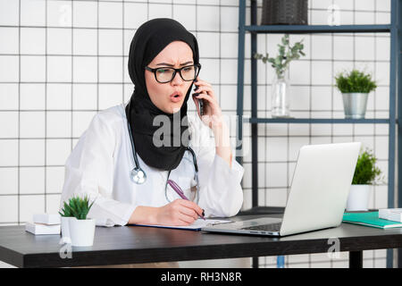 Weibliche verschleierte Wissenschaftler sitzen auf Ihrem Schreibtisch am Laptop auf der Suche öffnet sich ihr Mund mit entsetzten Gesicht Emotionen beim Gespräch mit einem Mobiltelefon Stockfoto