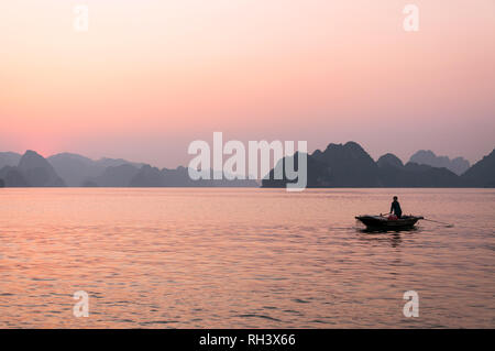 Eine vietnamesische Frau rudern Ihr holzboot mit Kalkstein Karst Inseln im Hintergrund bei Sonnenuntergang, Halong Bay, Vietnam Stockfoto
