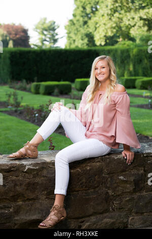 Eine attraktive blonde Haare kaukasischen High School senior posieren für ältere Bilder. Weibliche Teenager Portrait sitzen und Lächeln, draußen im Park in s Stockfoto