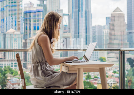 Junge Frau, die an einem Notebook arbeitet auf Ihrem Balkon mit Blick auf die Wolkenkratzer. Freelancer, remote Arbeiten, Arbeiten-von-zu-hause Stockfoto
