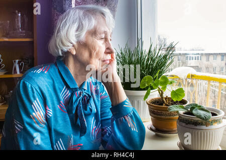 Alte, einsame Frau sitzt am Fenster in seinem Haus. Stockfoto