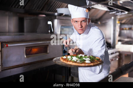 Jungen Koch in der Nähe von professionellen Backofen im Inneren des modernen Restaurant Küche, hält in seinen Händen Schaufel mit gekochtem Pizza Stockfoto