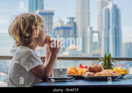 Der Junge ist beim Frühstück auf dem Balkon. Frühstück mit Kaffee Obst und Brot, Croissant auf einem Balkon vor dem Hintergrund der großen Stadt Stockfoto